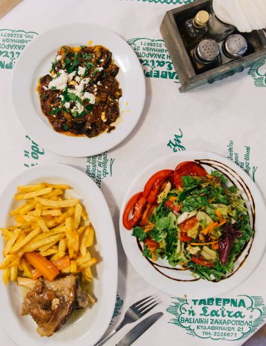 Plates of Greek food served on a table, french fries with lamb, green salad with tomatoe slices and smoked aubergines with feta cheese
