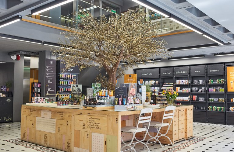A wooden stall with two chairs and an olive tree coming out of its centre, in a store selling beauty products.