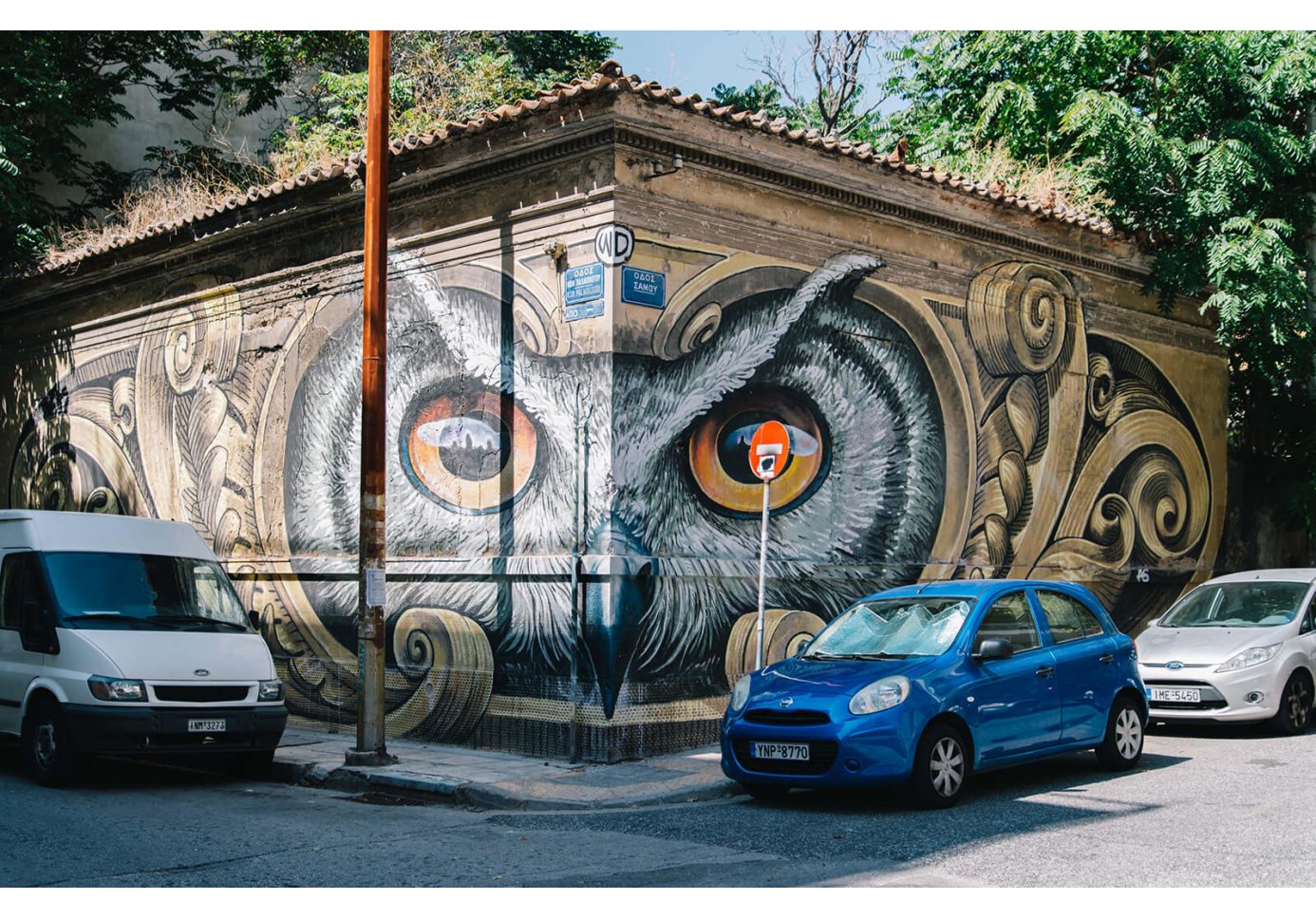 Graffiti of an owl's face on corner of two walls and 3 parked cars.