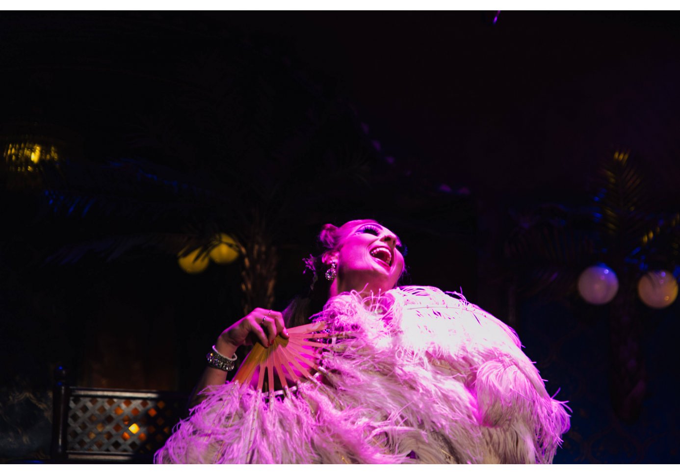 a drag performer laughing on stage holding a huge feathered hand fan.