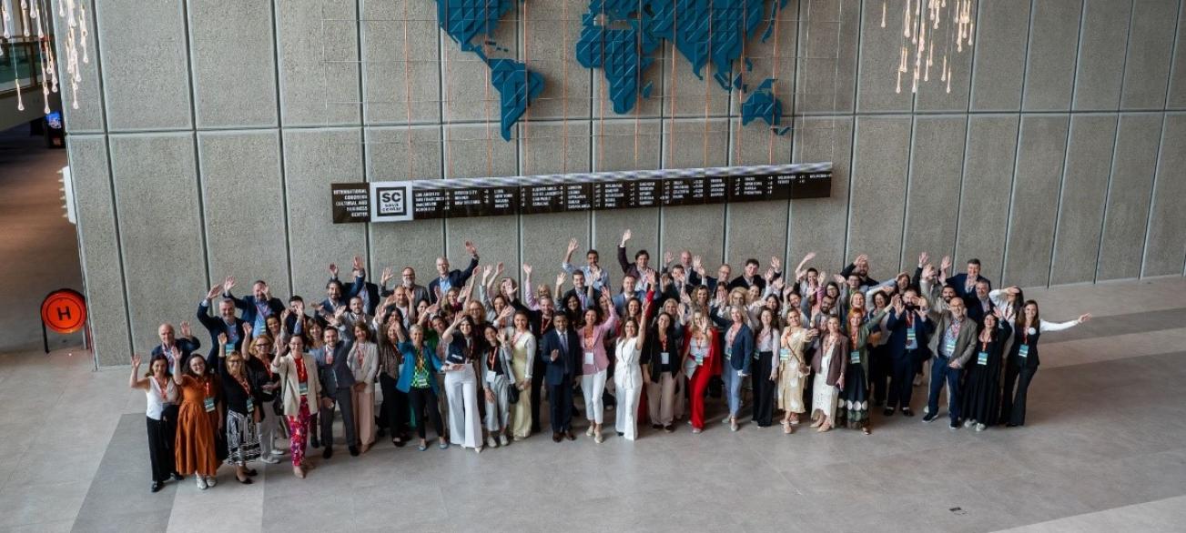 alt="A wide-angle shot of a diverse group of professionals at an international event. They are gathered for a group photo in a large atrium with stone-paneled walls, standing below a stylized blue world map."
