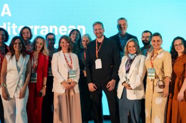 alt="A group of smiling professionals gathered on a stage for a photo, standing in front of a bright, light-blue digital screen. Most are wearing event lanyards."