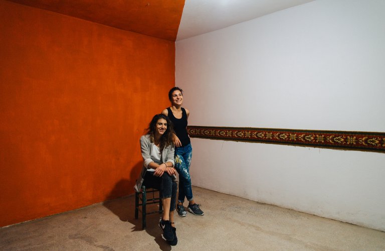 a woman sitting on a chair with another one leaning on her in front of a red and white wall