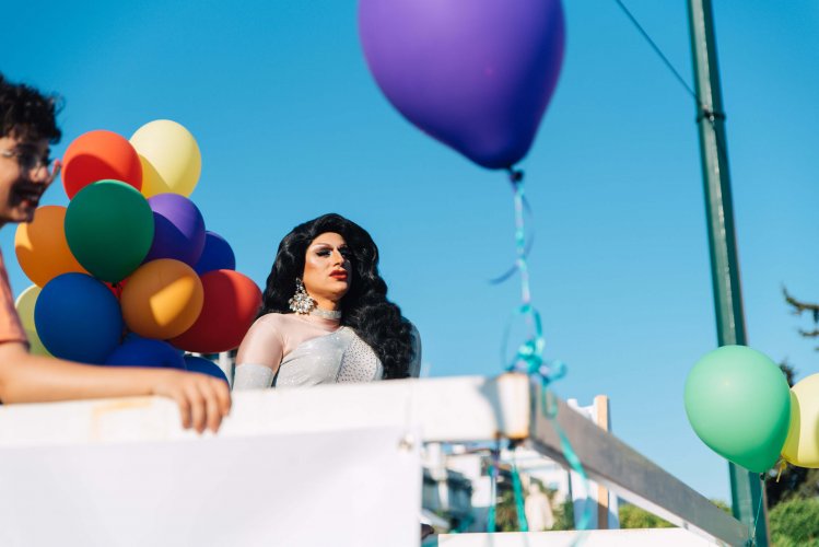a drag queen with pride coloured balloons posing on top of a truck.