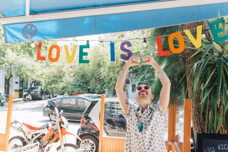 a man shaping a heart with his hands under a rainbow sign that reads "love is love" hanging from a blue tent.