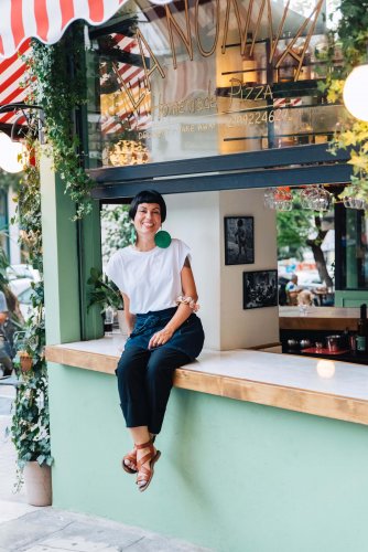 a woman sitting and smiling at the window of a pizza place, a sign reading "la nonna"