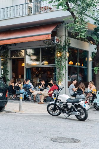 a corner shop, people sitting at tables outside, parked cars and motorbikes, sunny day