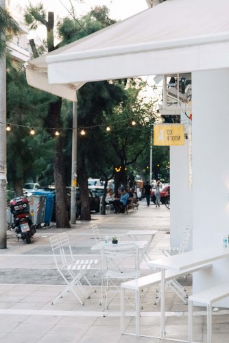 detail of a corner shop, empty white tables and chairs on the pavement, people, trees and cars at the background.