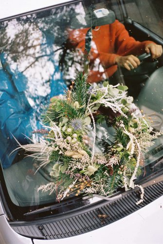 a flower wreath on a car bonnet, a man wearing an orange blouse is driving.