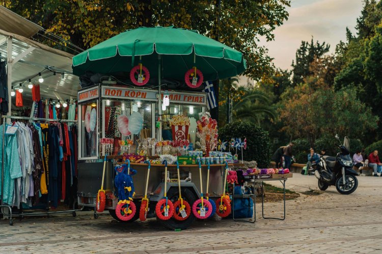 a kiosk in the park selling pop corn and toys, clothes hanging next to it, people sitting at the background. 