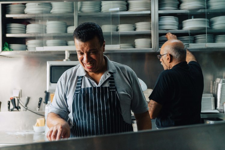 two men in a kitchen, one cooking, the other in front of shelves with stacked plates