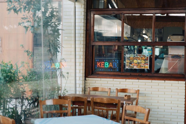 a shot of a restaurant, outdoor tables and window front, lit sign that reads "kebab" in red, green and blue letters
