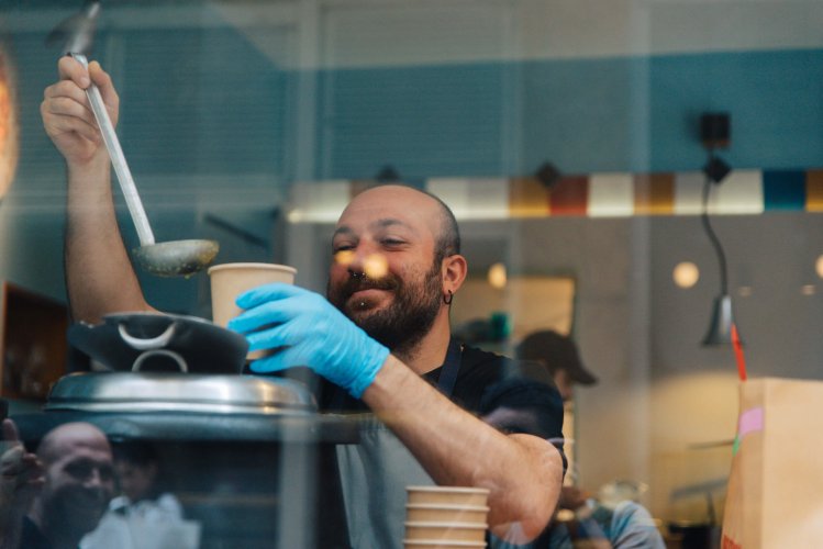 a man serving soup in a paper bowl, shot behind a window front