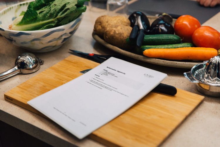 A wooden cutting board with a knife lying beside a recipe printed on paper, a plate of vegetables next to the cutting board.