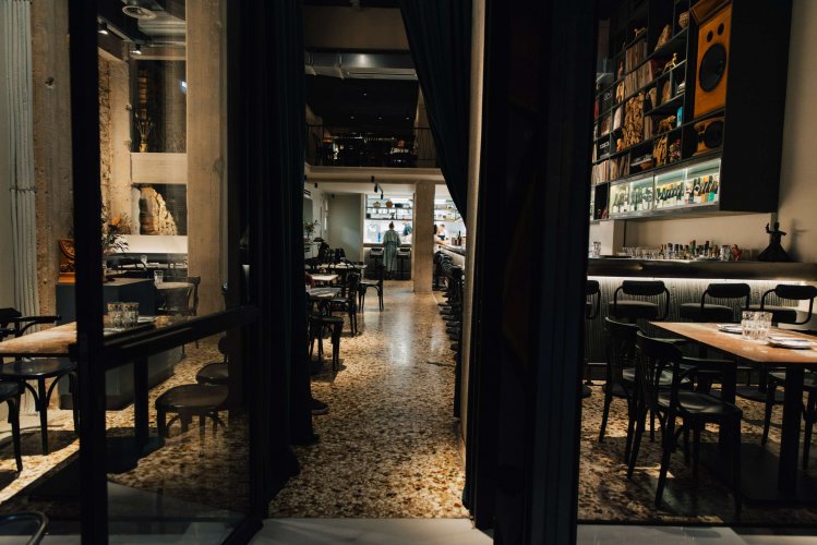 The interior of a restaurant with set tables and open kitchen viewed through a glass front door.