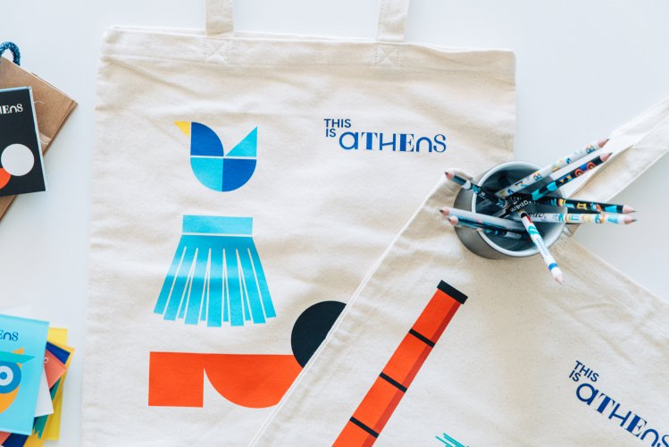 A tote bag with the text "This is Athens" in Greek lettering sits on a table next to a cup filled with pencils.
