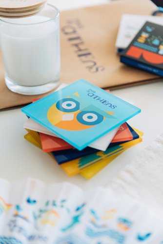A stack of colorful acrylic coasters with owl designs sits on a table. There is a glass of milk and a small fan next to the coasters. The text "Athens" is written above the owl design on the coasters.