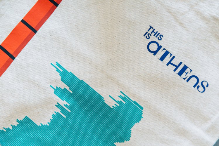 A close-up of a tote bag. The tote bag is canvas and appears to be natural colored. The text "this is athens" is printed on the front of the tote in a bold, dark font.