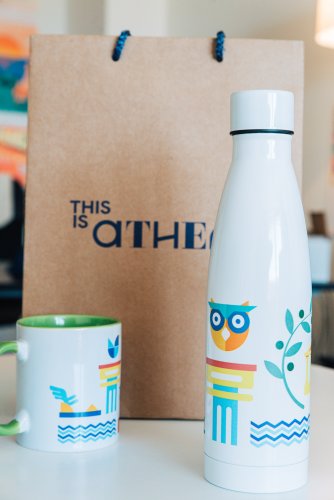 A metal water bottle and two ceramic mugs sit on a wooden table next to a brown cardboard takeout bag.