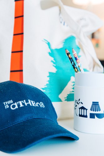 A blue baseball cap sits on a table next to a cup filled with pencils. A tote bag with the text "IS Athens" is in the background.