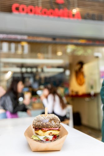 a burger, people eating at the backround and a sign that reads "cookoomela"