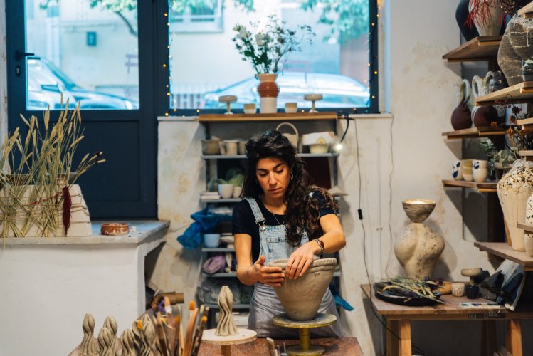 A woman in overalls shapes a clay pot on a wheel in a cosy pottery studio.