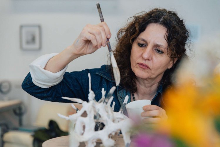 A woman with curly brown hair paints a ceramic sculpture while holding a cup in a studio.