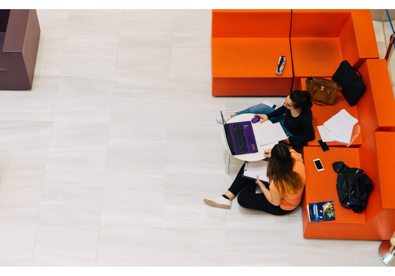 two girls sitting on the floor againsta orange chairs, working on their laptops