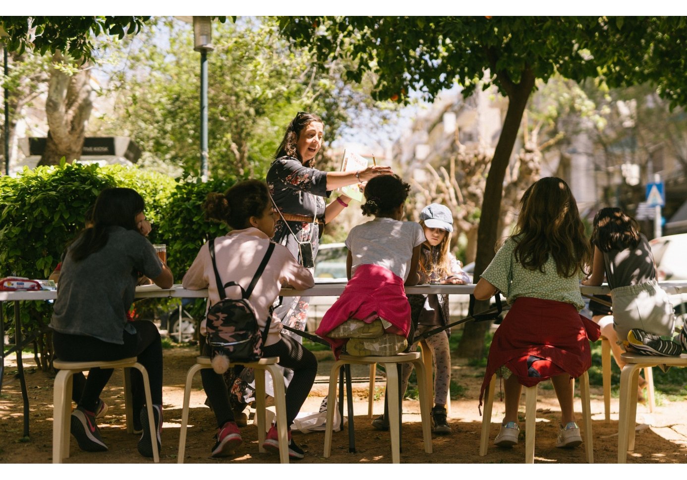 a woman directing kids who sit around a table at the park.