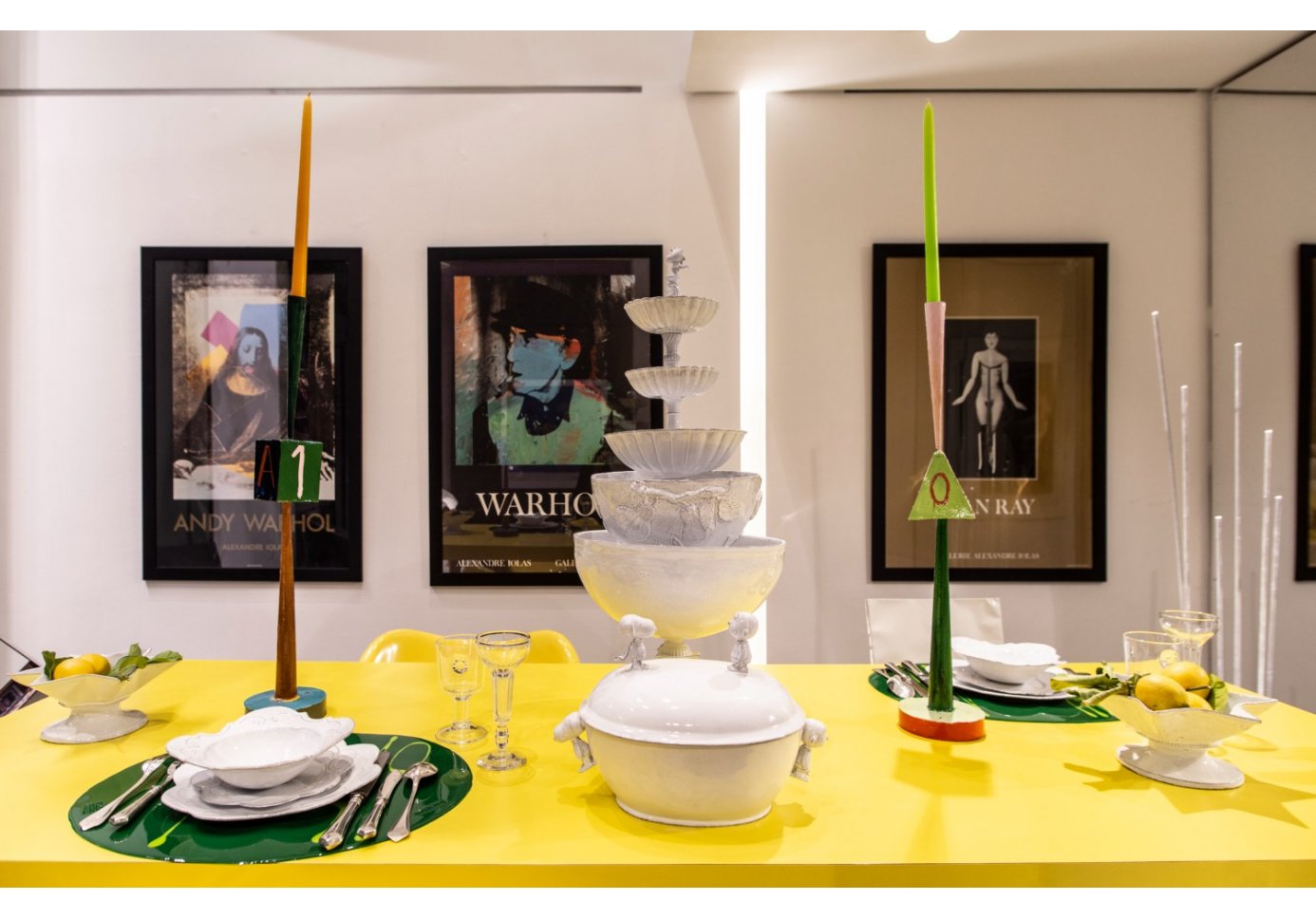 a yellow dining table, dishes and candles on top, three paintings on the wall behind. 