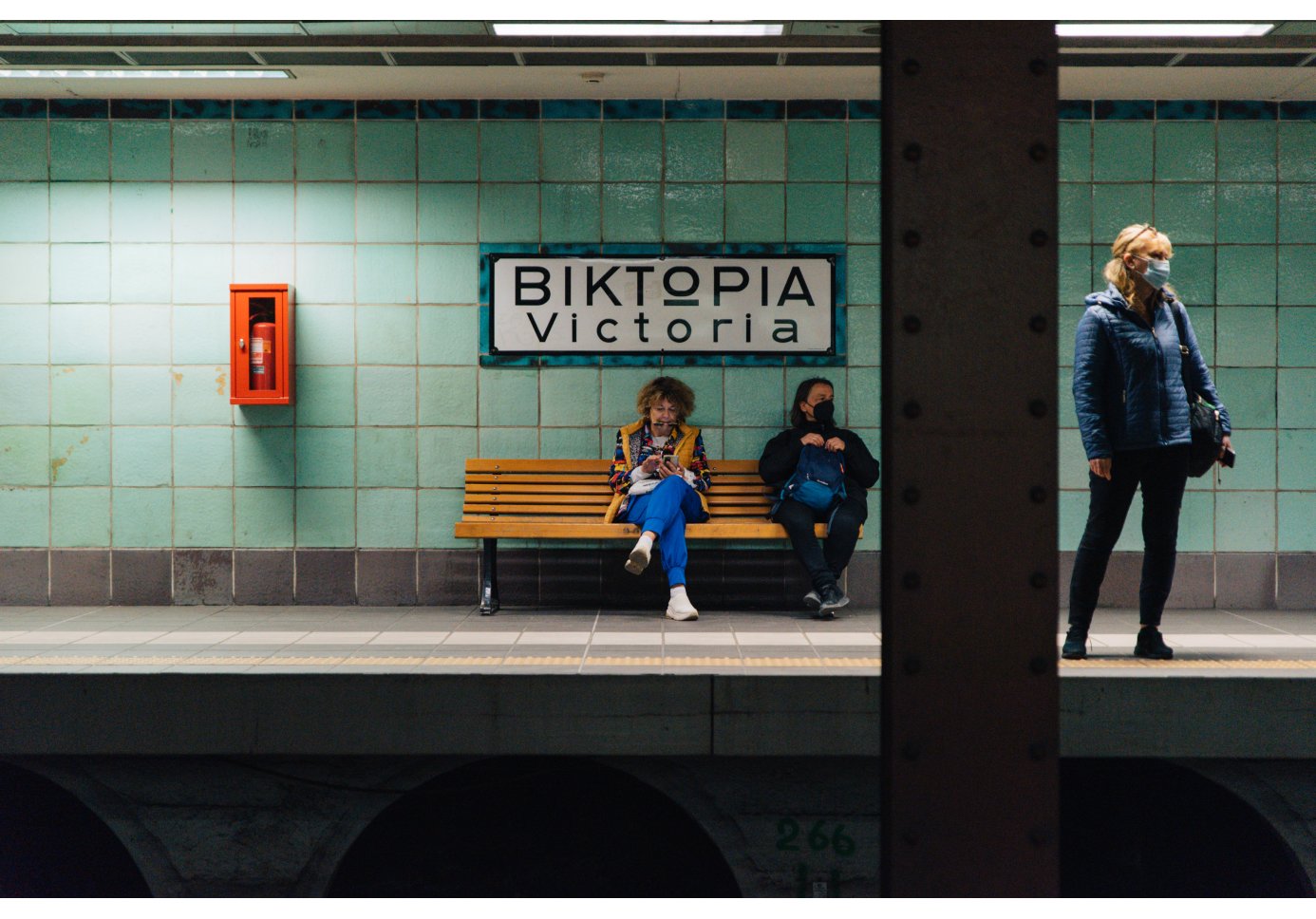 a metro platform with green tiles, two women sitting on a bench, one standing, a sign that reads "Victoria"