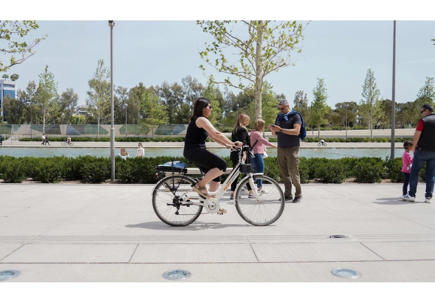 a woman in black biking in front of a man-made canal, people in the picture walking, trees