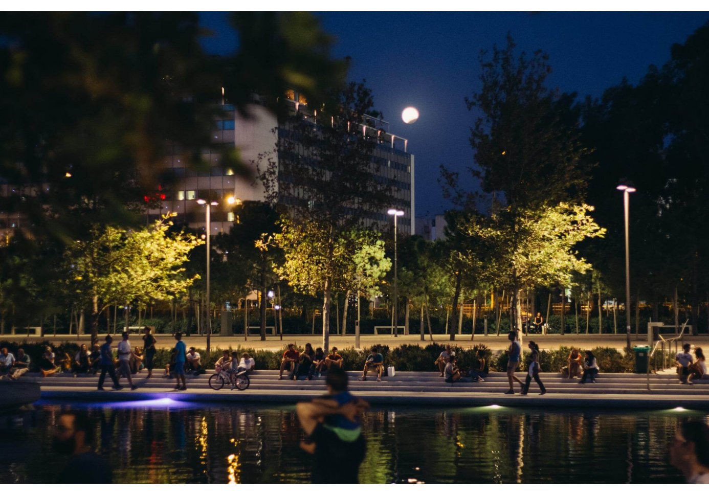 people sitting along a man-made canal in a park, night-time, full moon and an eclipse in the sky