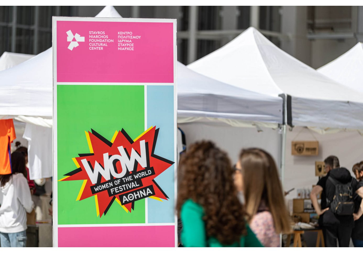 two women passing in front of a banner that reads "wow - women of the world festival athens", white tents in the background