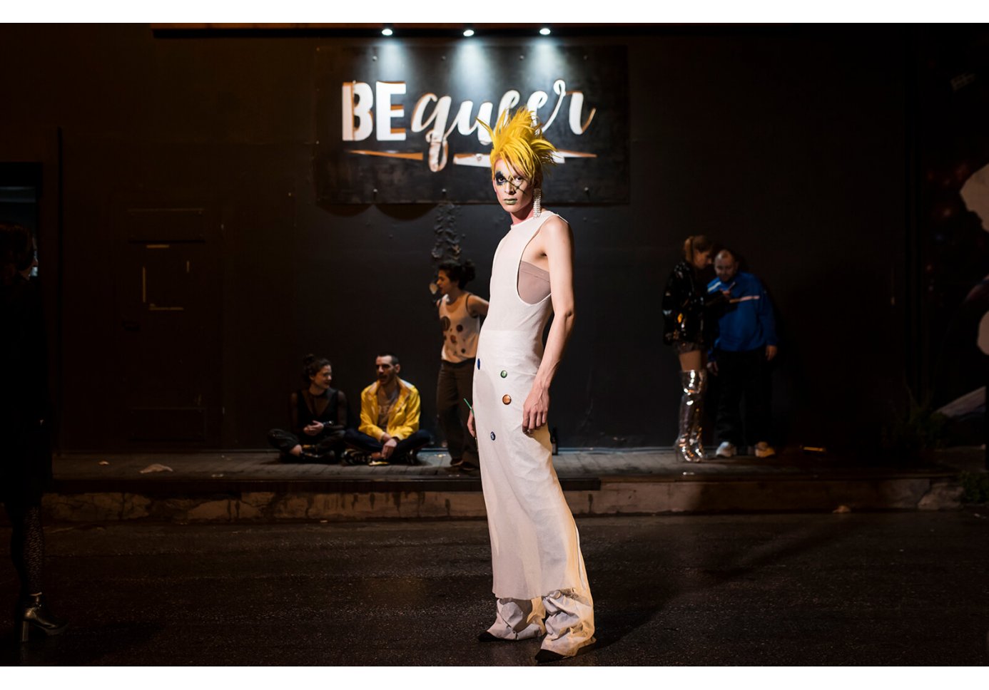 a man looking at the camera, other people sitting on the sidewalk outside of a nighclub